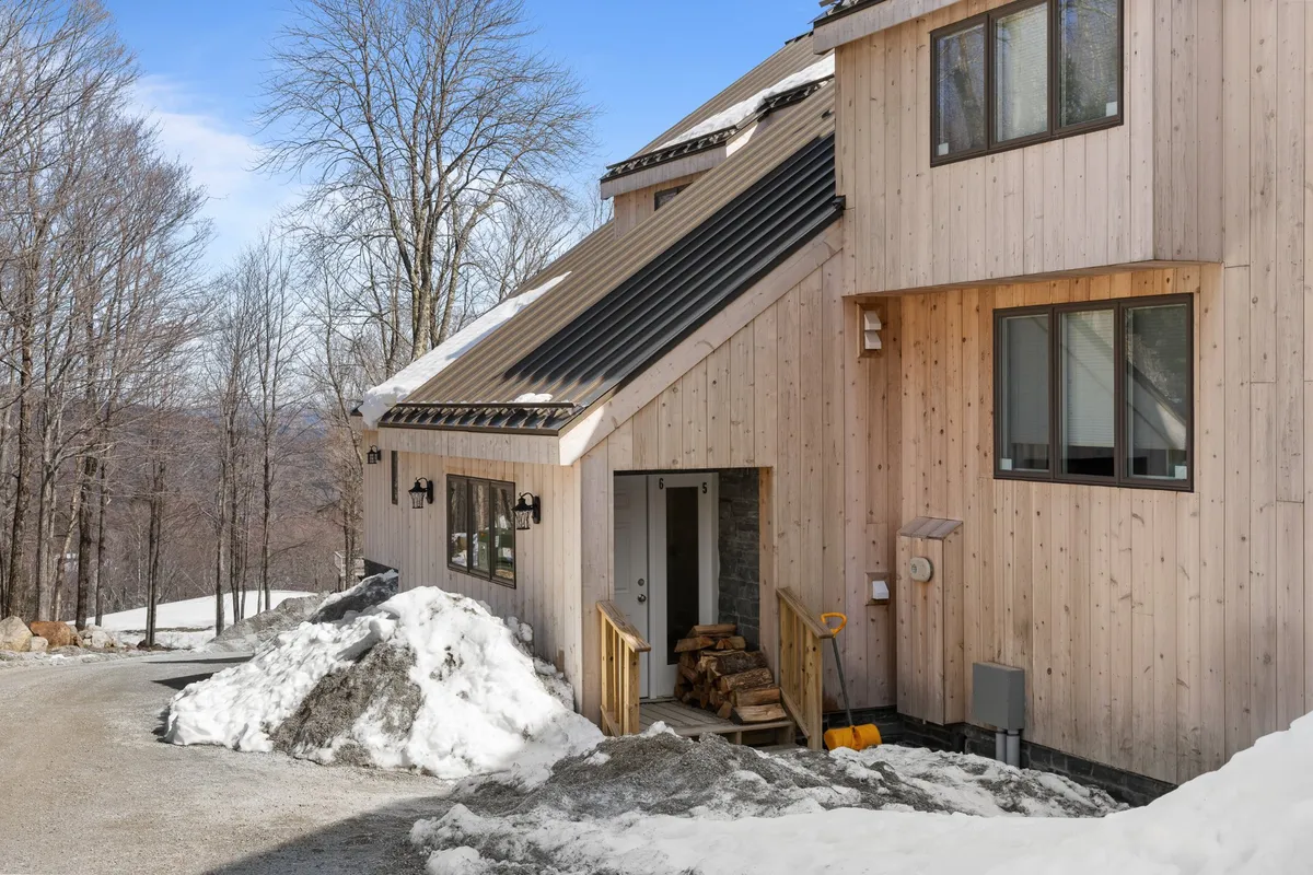 A cozy, welcoming exterior view of a wooden home nestled among trees, with a sloped roof and logs stacked by the front entrance. The setting is surrounded by natural beauty, suggesting a perfect home base for outdoor activities in all seasons.