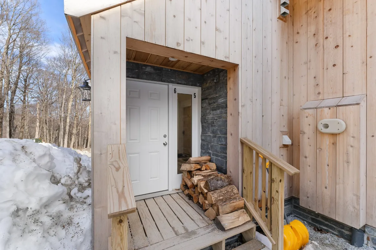 An exterior view of the condo's front door framed with a wooden railing and a neatly stacked pile of firewood. The scene feels welcoming and offers a glimpse of the natural mountain environment, suggesting direct outdoor access for adventures.