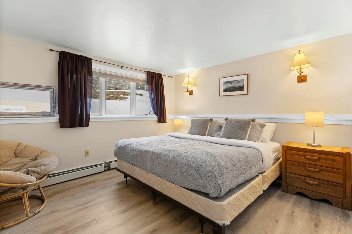 This image displays a serene bedroom with a comfortable bed, a window letting in gentle natural light, an armchair, a nightstand, and tasteful artwork. The scene is inviting and tranquil, perfectly representing a restful retreat.
