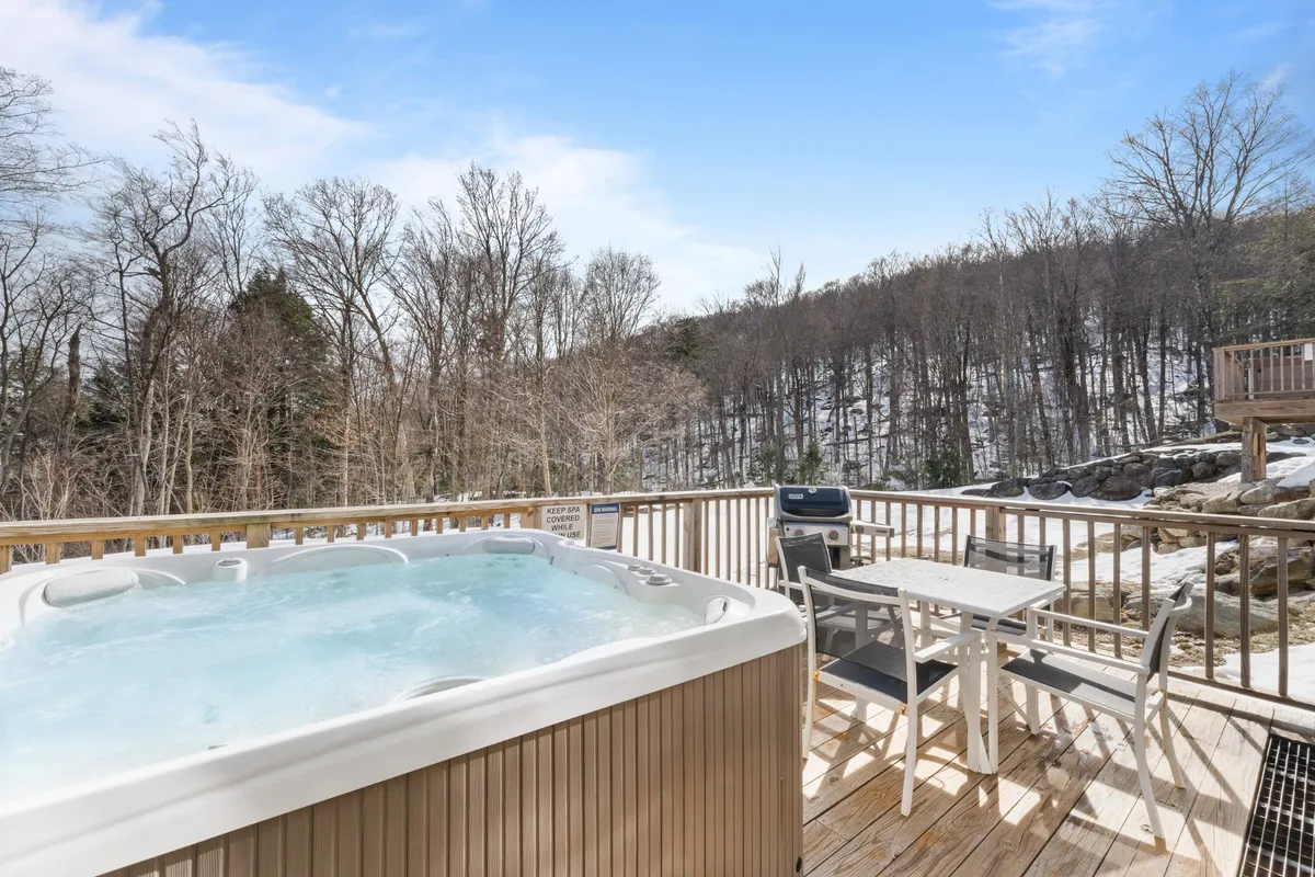 A tranquil outdoor setting showing a hot tub situated on a wooden deck surrounded by trees. The space is clearly set up for both relaxation (hot tub) and social moments (BBQ grill area), with nature providing a peaceful backdrop for all-season enjoyment.