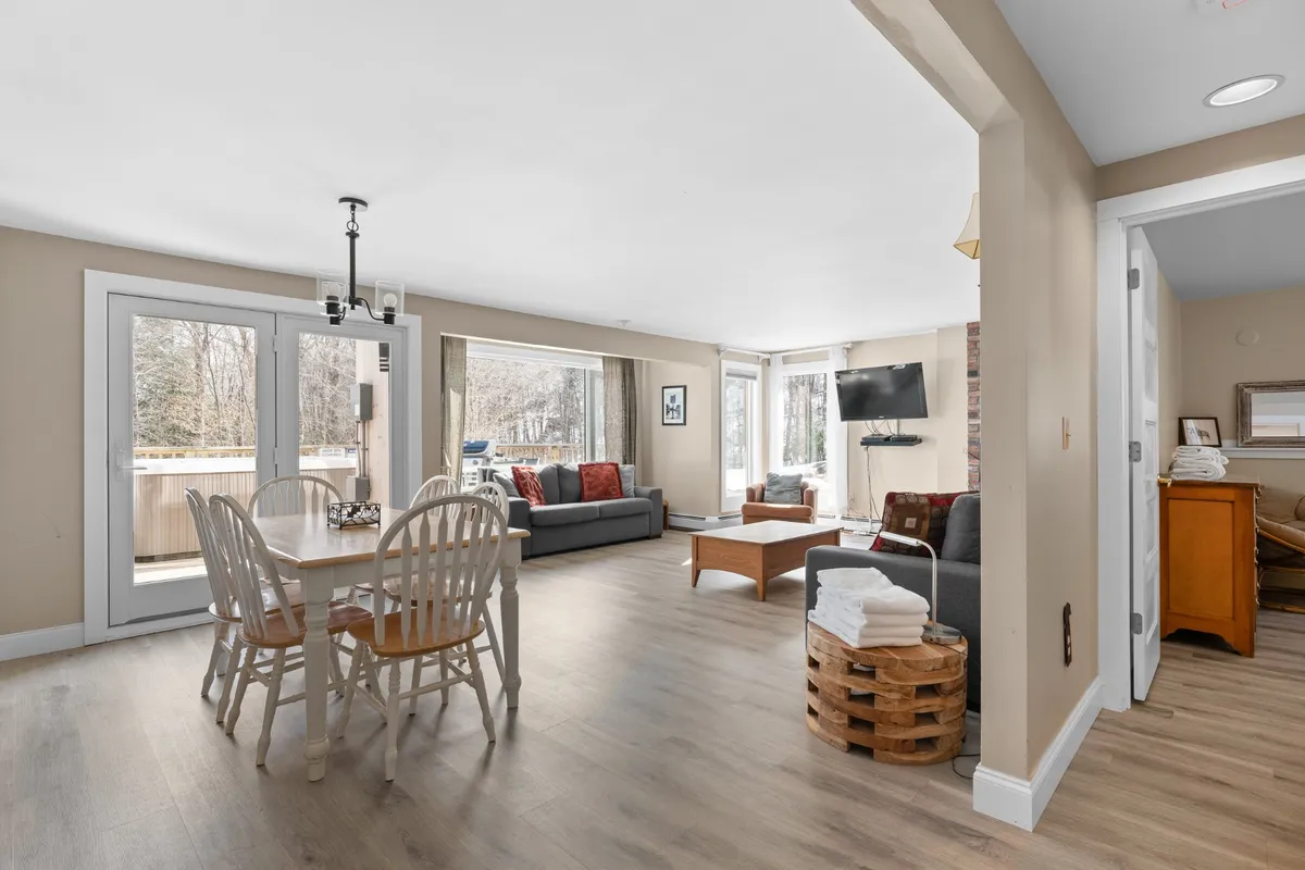 The image features a bright, inviting living space with a cozy sofa, a stylish dining table set for meals, and expansive windows flooding the room with natural Vermont sunlight. The television is perfectly positioned for relaxed movie nights, while the overall layout fosters socializing and family togetherness.