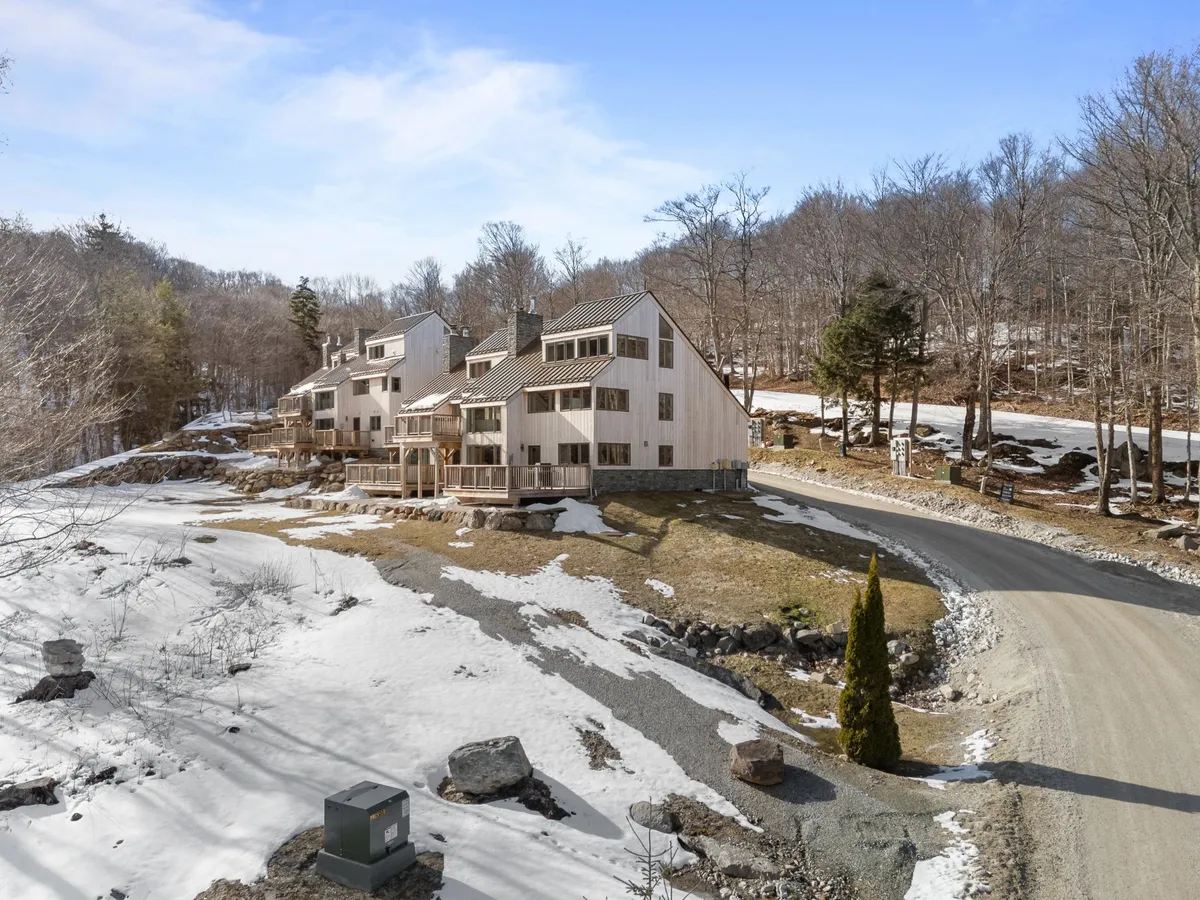 This exterior photo features modern architecture set against a snow-blanketed landscape, framed by a tree line and a curved driveway. The setting feels serene yet inviting, suggesting easy access to both natural beauty and the amenities of Killington Village.