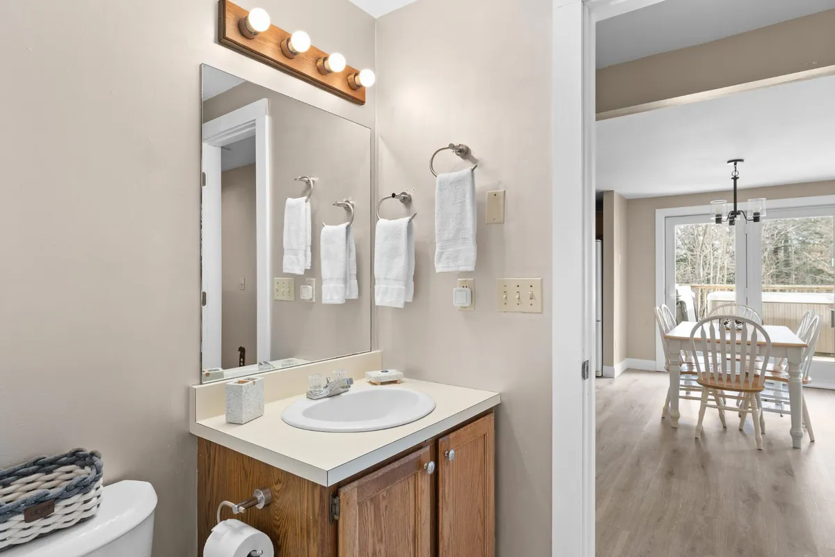 A cozy bathroom scene highlights a well-lit sink and mirror, wooden cabinetry, fresh towels hanging on hooks, and a glimpse of the dining area through an open doorway. The setting feels clean, welcoming, and thoughtfully appointed.