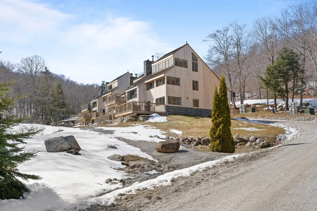 A modern house sits on a snow-blanketed property with a curved driveway leading up to the entrance. The architecture is clean and inviting, framed by a natural landscape, and hints at easy access to outdoor activities like skiing and hiking.