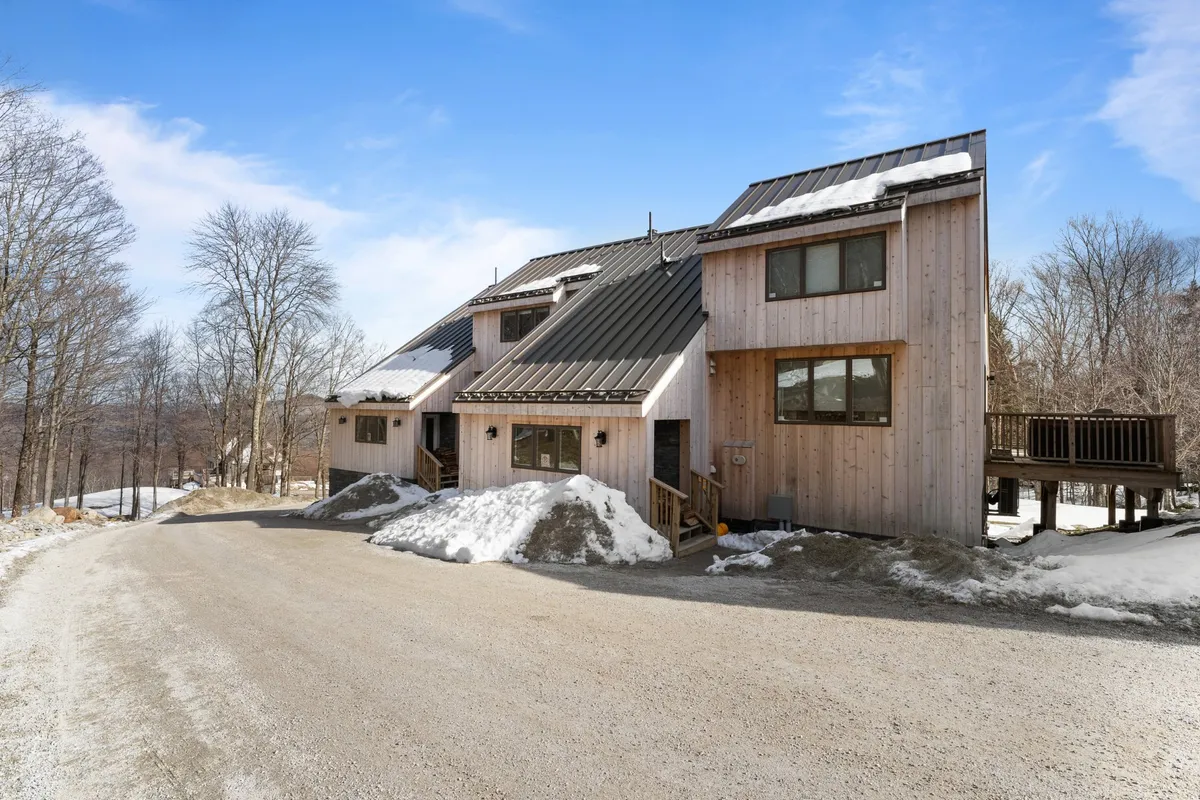 The wooden house is shown with a sloped metal roof surrounded by trees and set against a clear blue sky. The inviting deck and open space highlight the possibilities of gathering outside, whether for snow play in winter or warm-weather relaxation.
