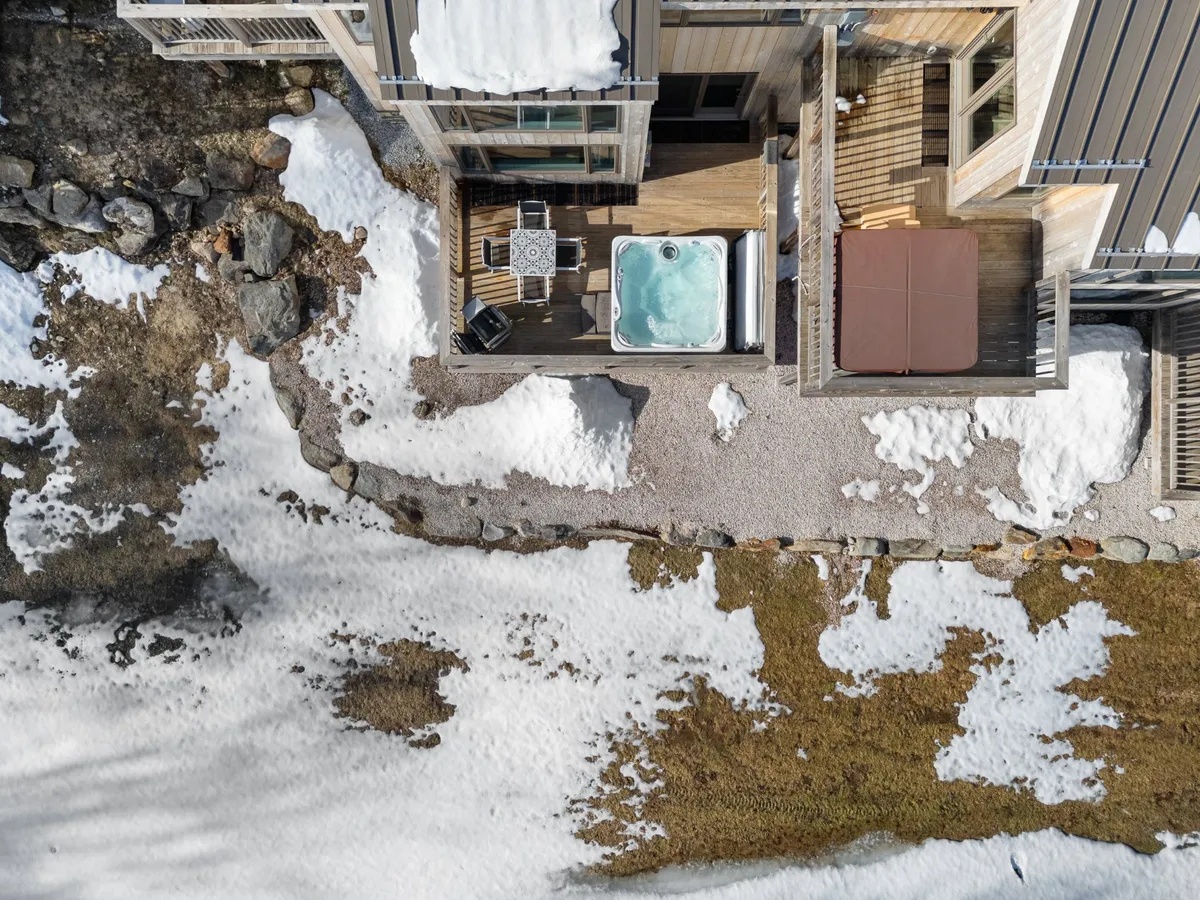 An inviting outdoor hot tub set amidst a tranquil seating area, with the ground blanketed in snow and natural rock formations nearby. The setup is ideal for relaxing while taking in the surrounding seasonal scenery, and the space can accommodate gatherings for barbecues or stargazing.