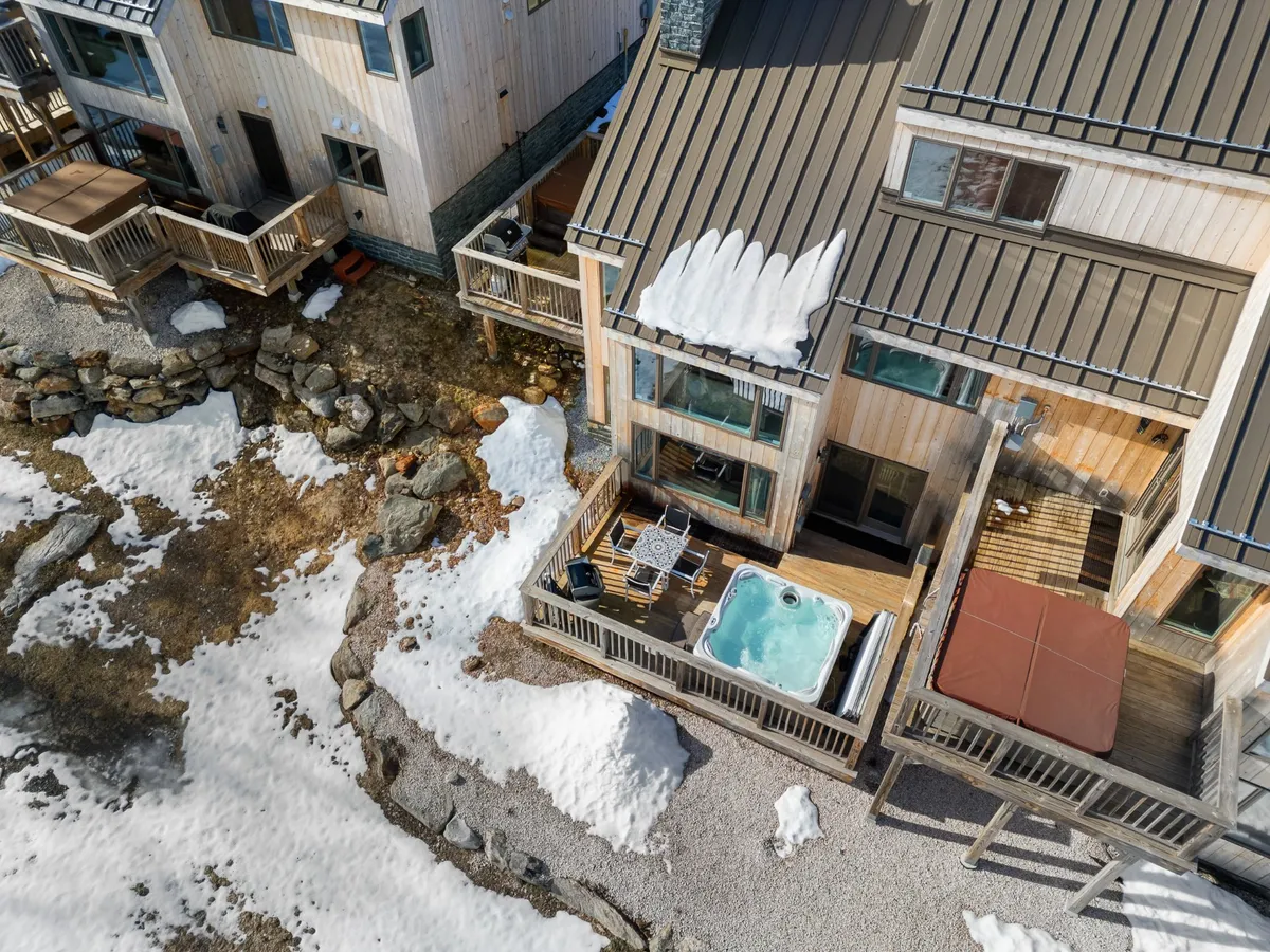 A hot tub sits on a deck surrounded by snow, with outdoor seating, rocky landscaping, and distant trees. The area exudes relaxation and togetherness, perfect after a day of family adventure in the Vermont wilderness.