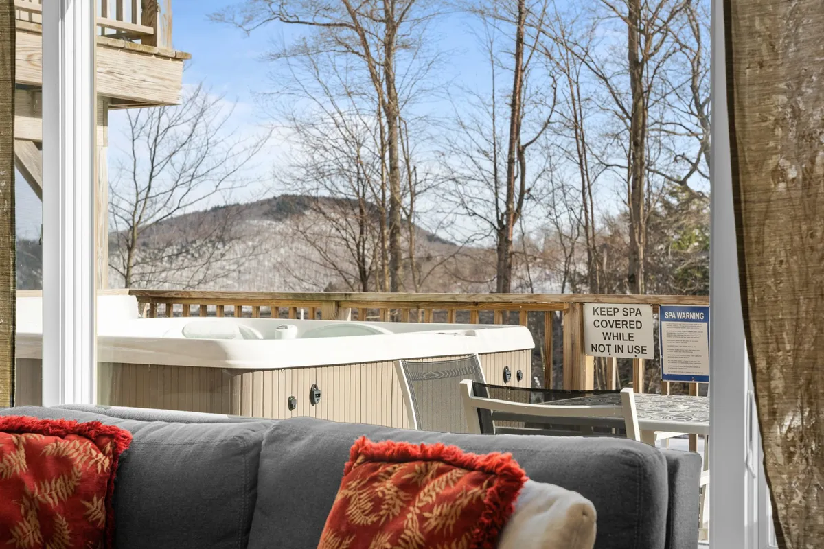 A cozy indoor space featuring a hot tub, plush sofa with red pillows, and expansive mountain views through the windows. The inviting setting combines elements of relaxation and socializing, with the outdoors clearly visible to evoke a connection to the surrounding landscape.