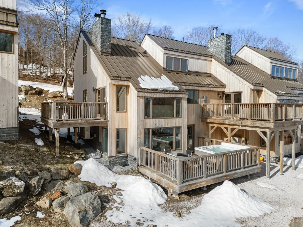 An exterior perspective of the home’s deck featuring a hot tub, wooden siding, and large windows. The deck looks out onto peaceful surroundings and is portrayed as a place for quiet moments or group gatherings, with the hot tub and deck as focal points.