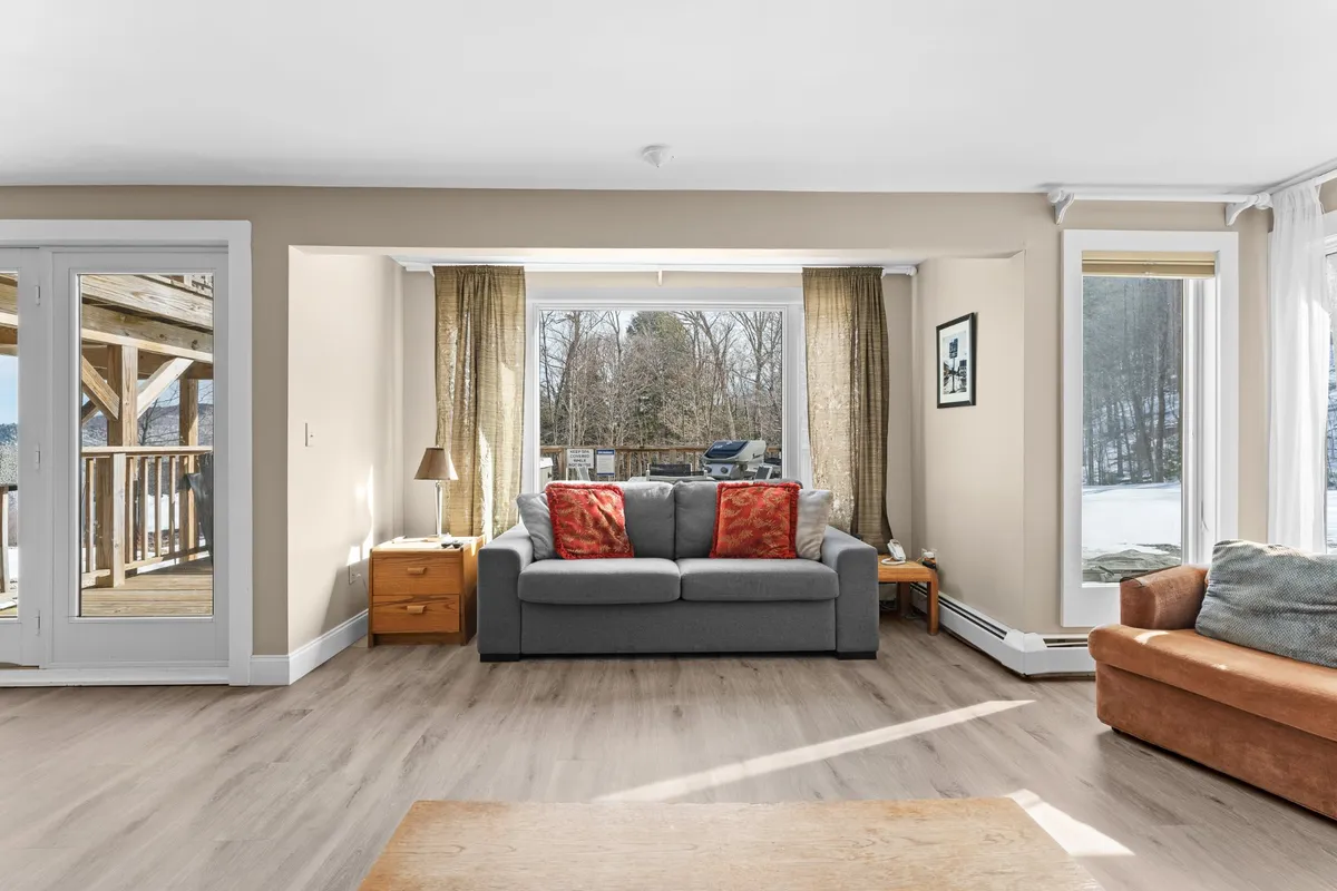 A cozy living room featuring a plush sofa with vibrant red pillows, a wooden table, an inviting armchair, and expansive windows that frame stunning mountain views. The space is bathed in natural light, creating an ideal setting for relaxation, socializing, or simply enjoying the picturesque surroundings.